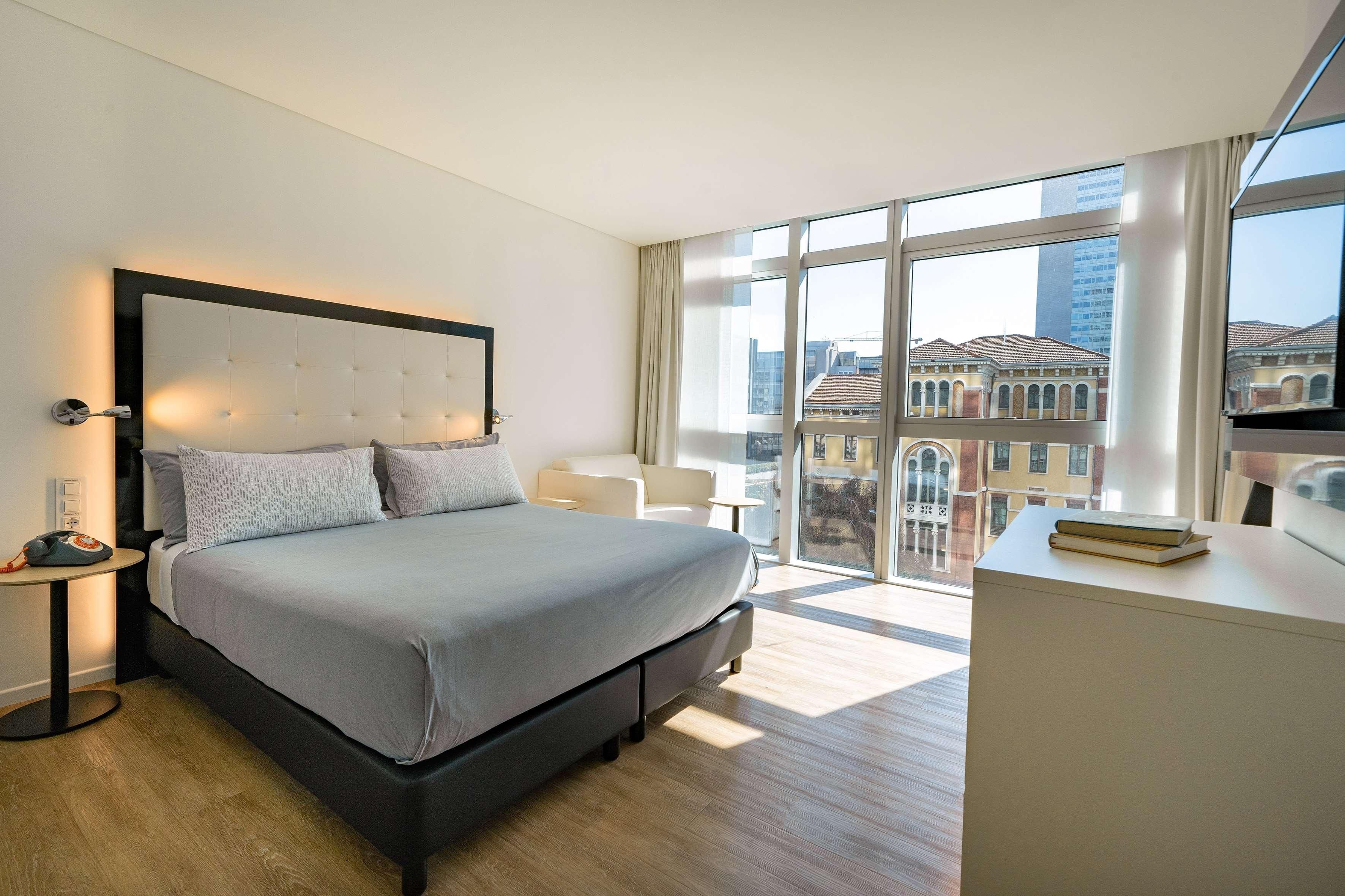 A modern hotel room with a large bed, a bedside table, a desk, and a view of the city through large windows.