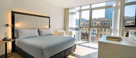A modern hotel room with a large bed, a bedside table, a desk, and a view of the city through large windows.