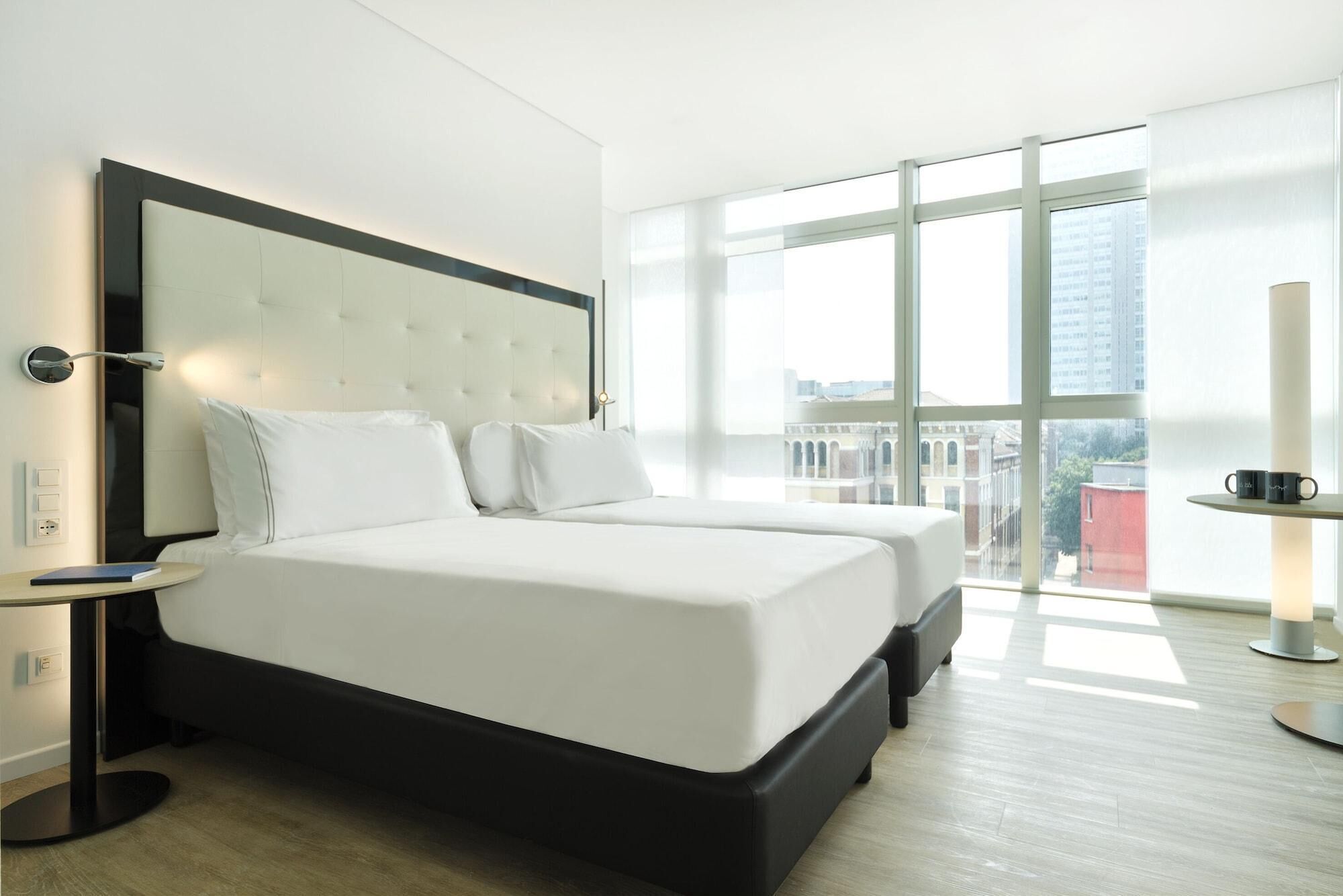 A modern hotel room with a large bed, a bedside table, a lamp, and a view of the city through large windows.