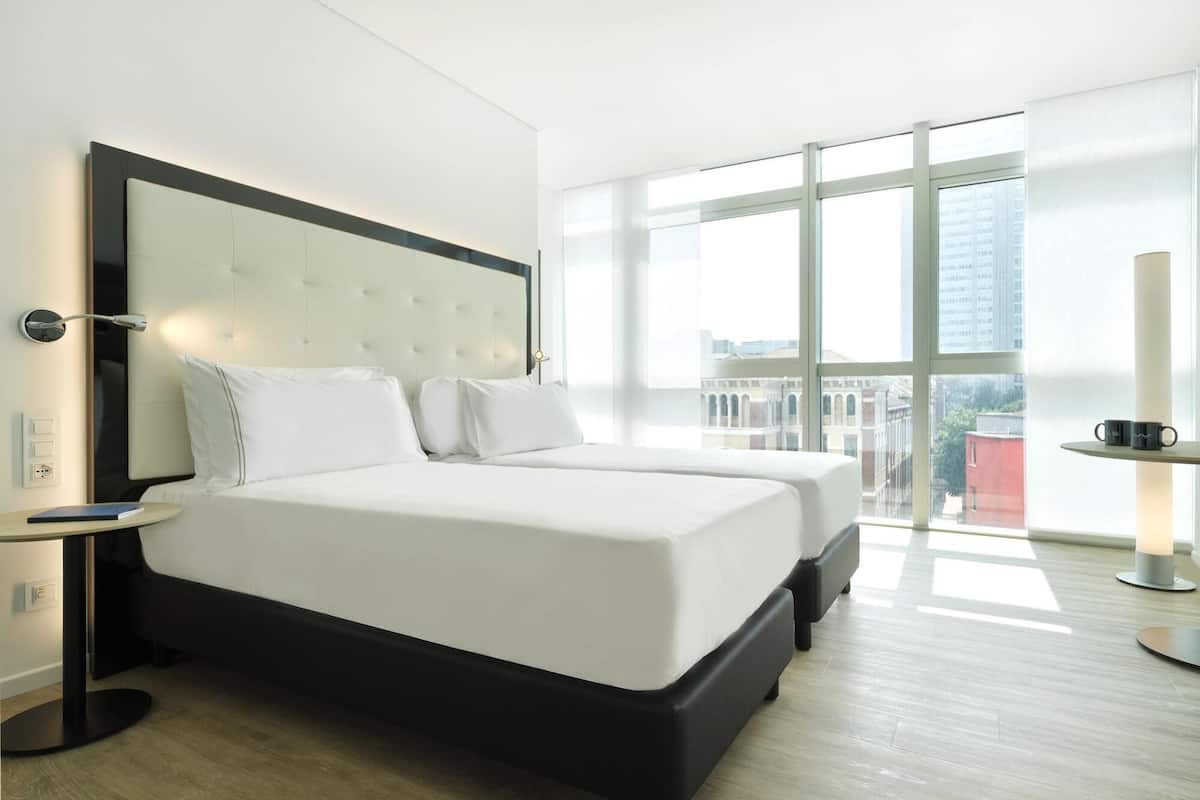 A modern hotel room with a large bed, a nightstand, a bedside table, and a view of the city through large windows.