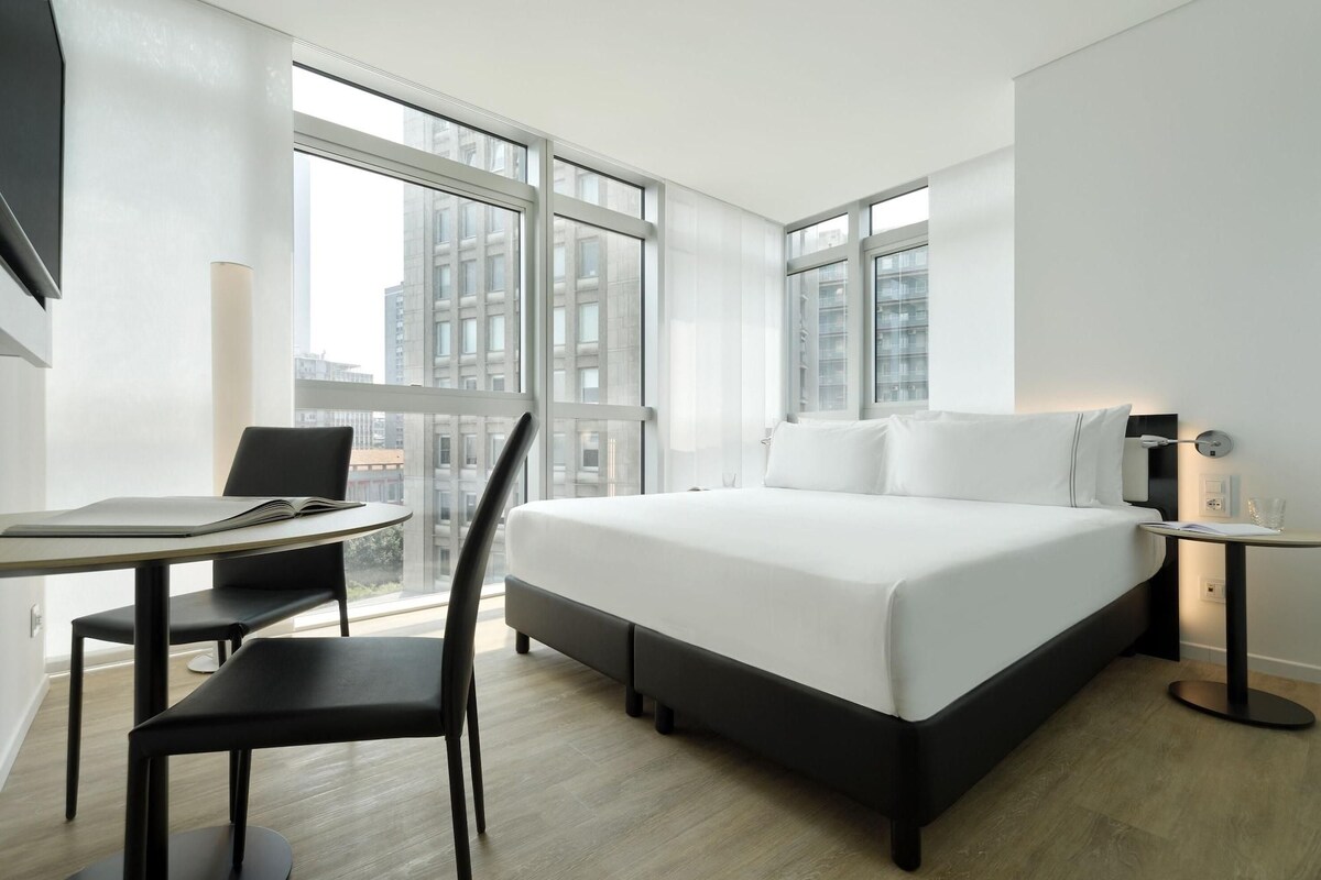 A modern hotel room with a large bed, a desk with a chair, and a window offering a city view.