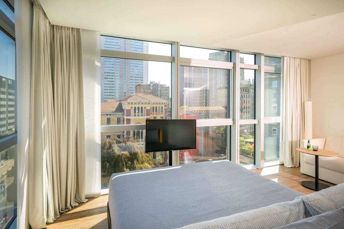 A modern hotel room with a large bed, a TV on a stand, a small table, and a view of the city through large windows.