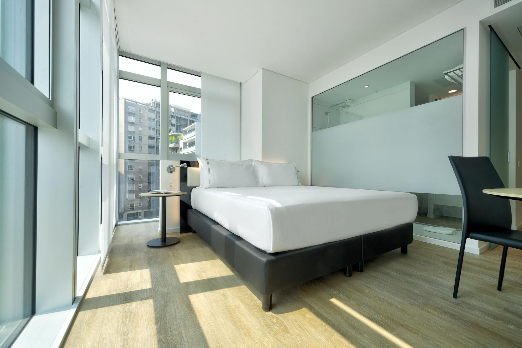 A modern hotel room with a large bed, a small bedside table, a chair, and a large window offering a city view.
