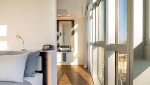 Una habitación de hotel moderna con cama, mesita de noche y vistas a la ciudad a través de amplias ventanas.