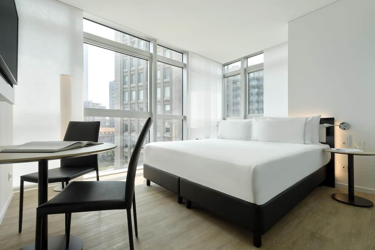 A modern hotel room with a large bed, a desk with a chair, and a window offering a city view.