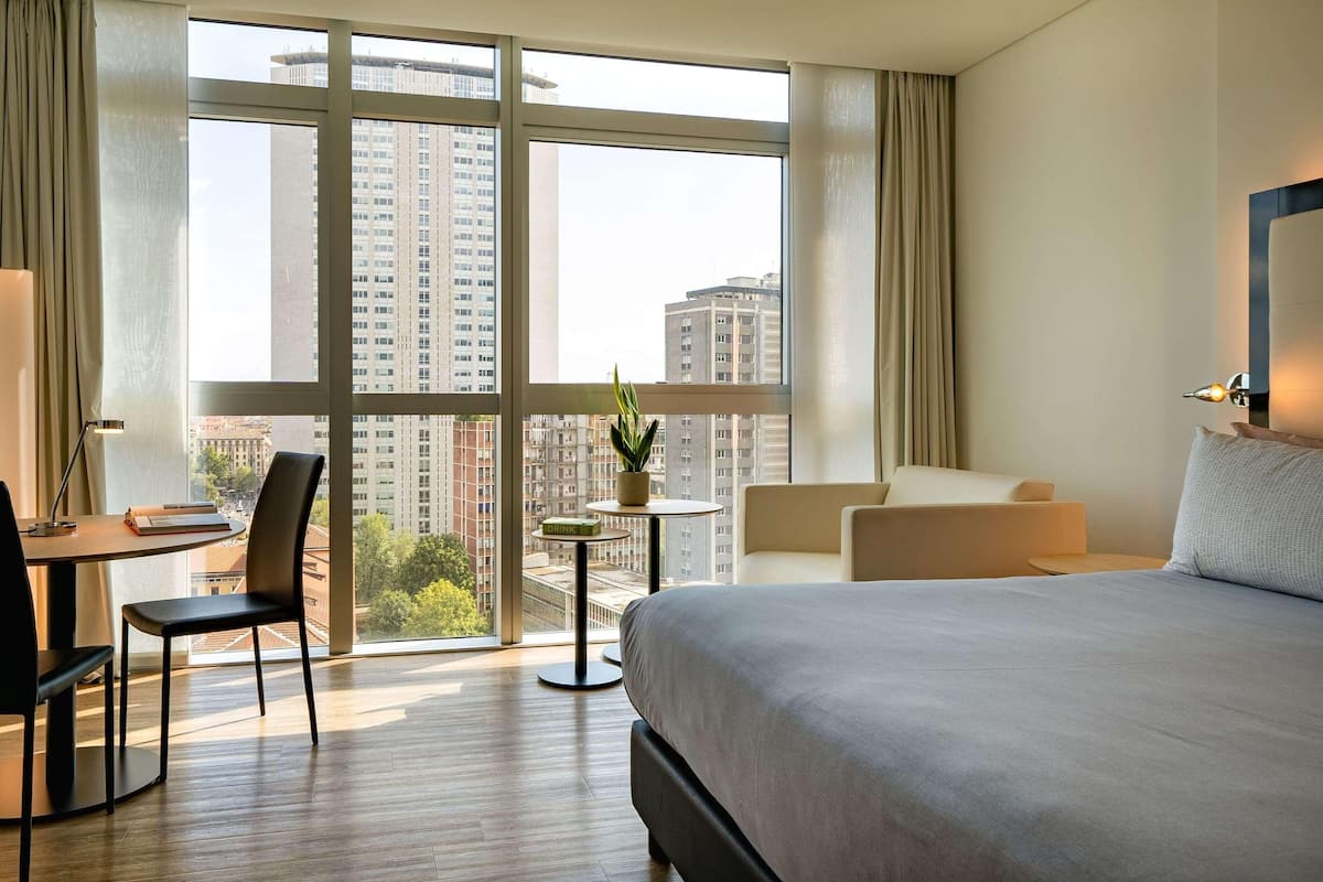 A hotel room with a large bed, a desk with a chair, a small table with a plant, and a view of tall buildings through large windows.