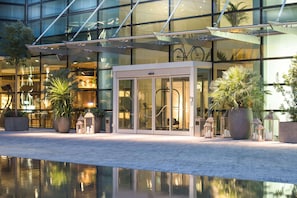 Una entrada de hotel moderna con puertas de vidrio, plantas en macetas y una piscina reflectante.