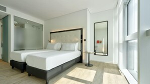 Quarto de hotel moderno com duas camas, um espelho grande e vista para a cidade pela janela.