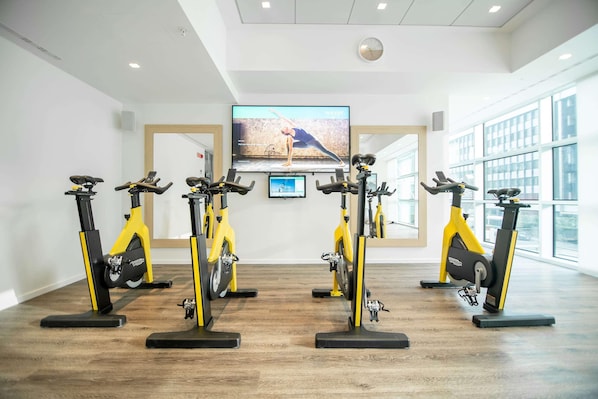 A gym with four stationary bikes arranged in a row, a large TV screen displaying a yoga video, and large windows providing natural light. - Innside Milano Torre Galfa (Milan)