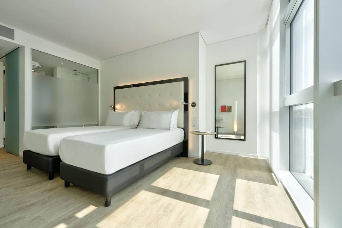 A modern hotel room with two beds, a large mirror, and a view of the city.