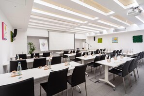 Une salle de conférence avec des tables blanches, des chaises noires et un écran de projection.