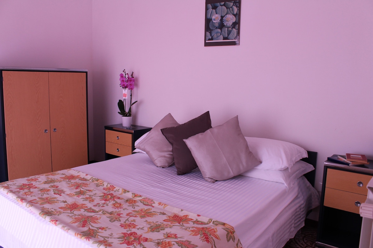 A neatly made bed with floral bedding, a nightstand with a book, and a vase with flowers.