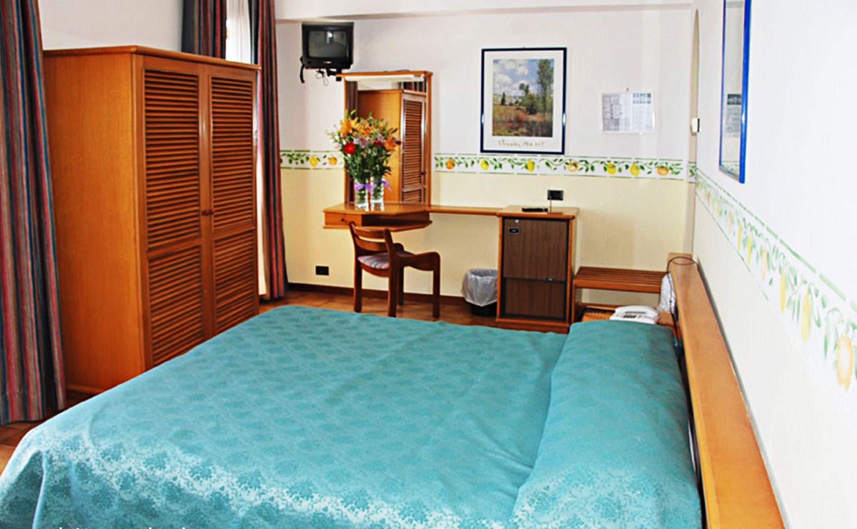 Quarto de hotel com cama, secretária, cadeira, guarda-roupa e televisão.