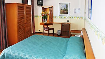 Quarto de hotel com cama, secretária, cadeira, guarda-roupa e televisão.