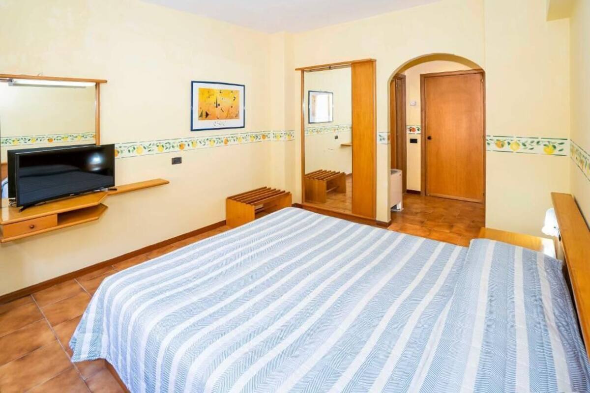 A bedroom with a bed, a TV on a stand, a mirror, and a door leading to another room.