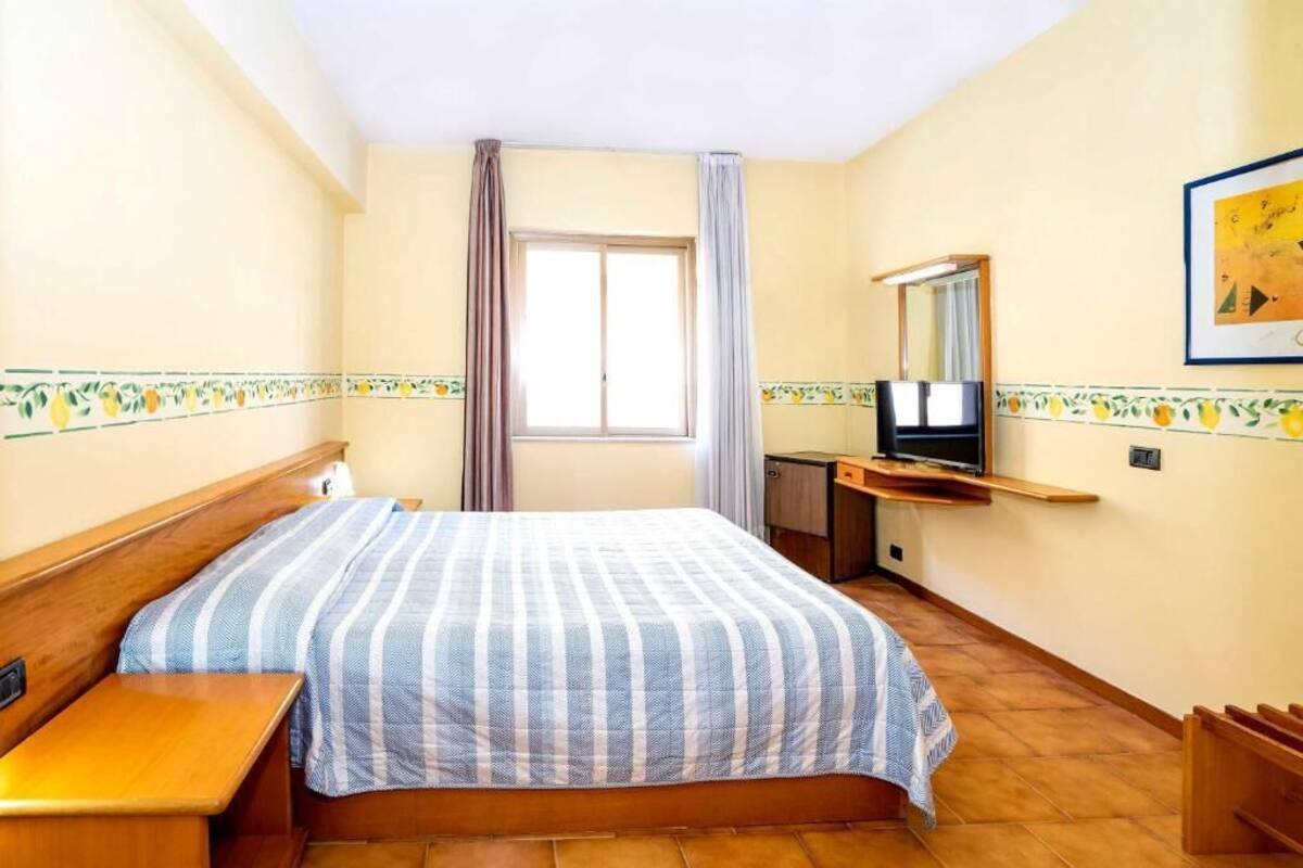 A bedroom with a bed, a wooden nightstand, a TV on a stand, a window with curtains, and a wall with a fruit pattern.
