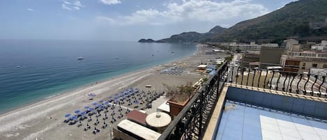 Una playa con sombrillas y tumbonas, cielo despejado y vistas al mar y colinas al fondo.