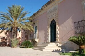 A building with a pink facade, a central arched doorway, and a staircase leading to the entrance. - Villa Principe Di Belmonte (Modica)