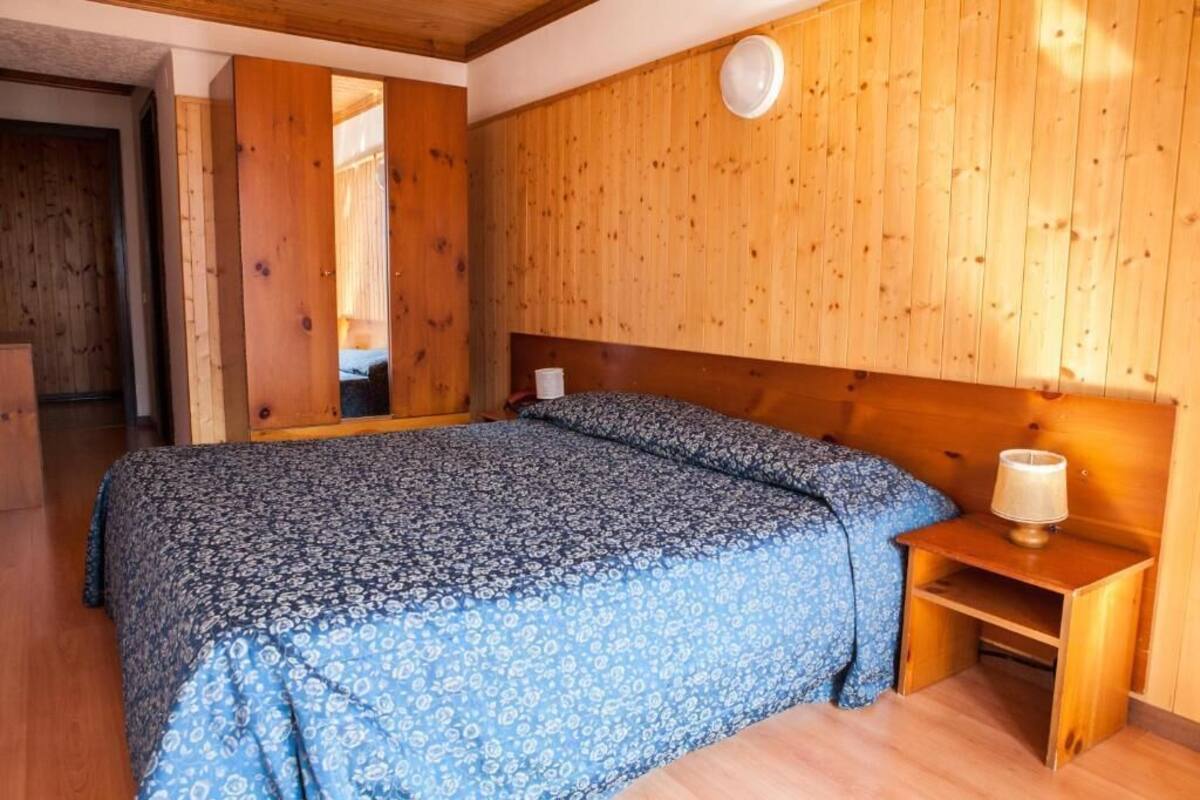 A bedroom with a wooden headboard, a bed with patterned bedding, a bedside table with a lamp, and a mirror on the wall.