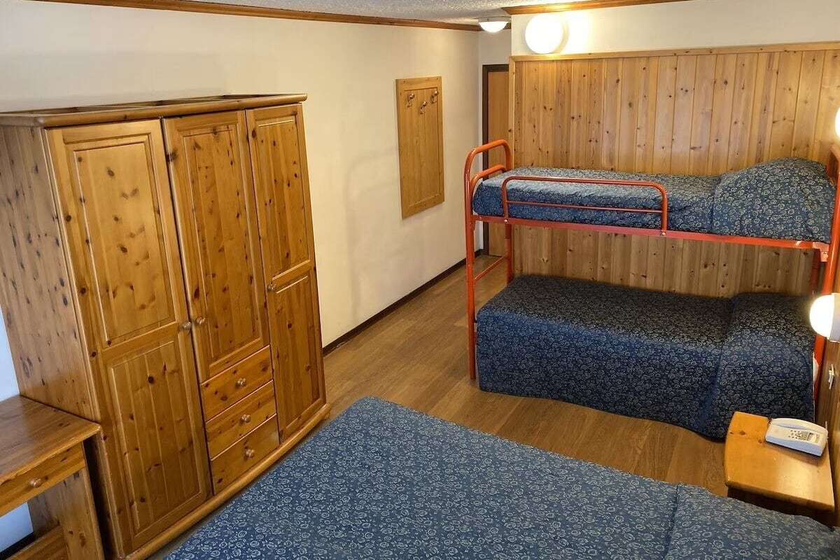 A bunk bed with a patterned mattress, a wooden wardrobe, and a small table with a lamp.