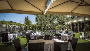 Un espace de restauration en plein air, avec des tables et des chaises placées sous de grands parasols, entouré de verdure et d’arbres.