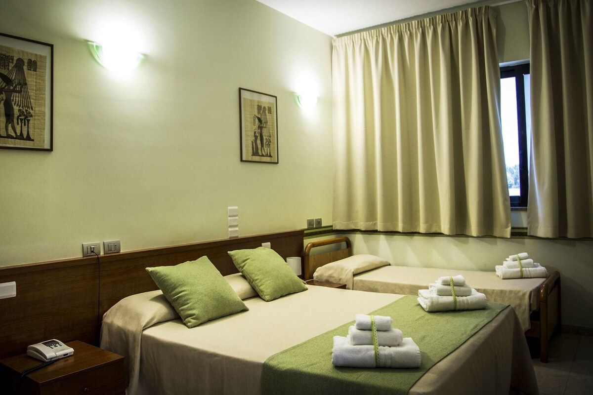 A hotel room with two beds, a wooden headboard, green pillows, a bedside table with a phone, and a window with curtains.