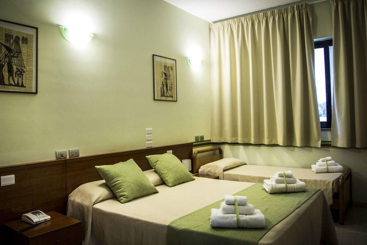 Une chambre d’hôtel avec deux lits, une tête de lit en bois, des oreillers verts et une fenêtre avec des rideaux.