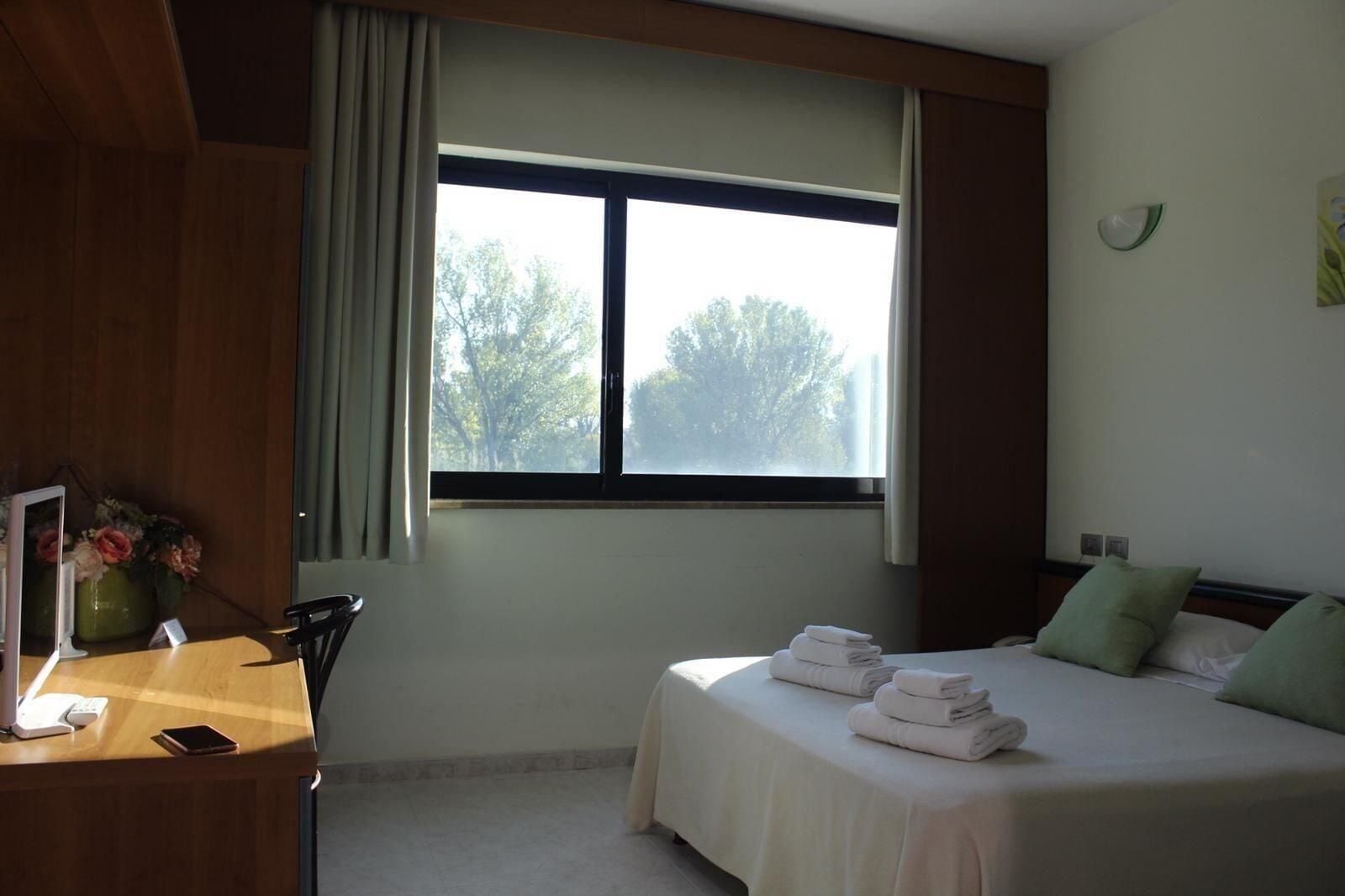 Una camera d'albergo con un letto, una scrivania con telefono e tablet, una finestra con tende e vista sugli alberi.