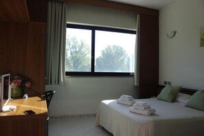 Une chambre d’hôtel avec un lit, un bureau doté d’un téléphone et d’une tablette, une fenêtre avec des rideaux et une vue sur les arbres à l’extérieur.