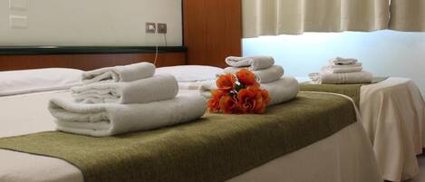 A hotel room with two beds, each with neatly folded white towels and a green throw blanket. A bouquet of orange flowers is placed on the bed between the beds.