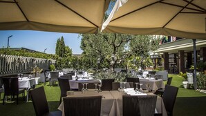 Un espace de restauration en plein air, avec des tables et des chaises placées sous de grands parasols, entouré de verdure et d’arbres.