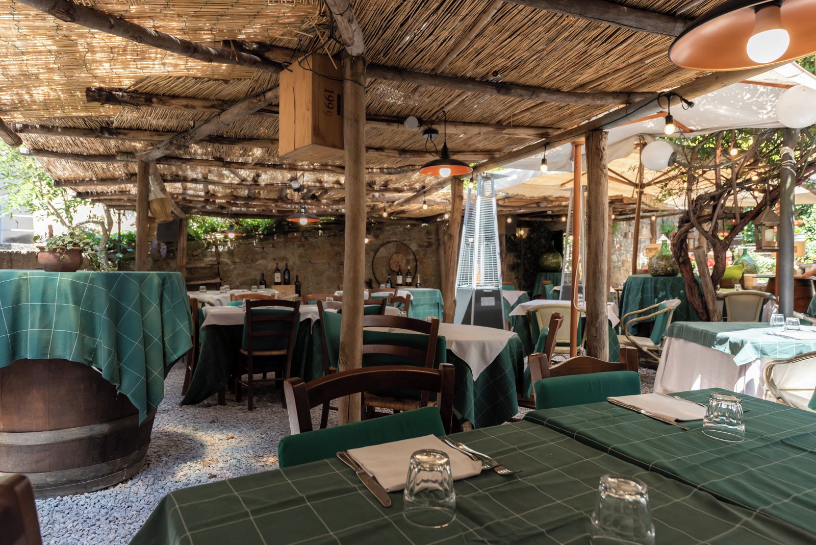 Ein Restaurant im Freien mit Tischen, die mit grünen Tischdecken bedeckt sind, Holzstühlen und hängenden Pendelleuchten.