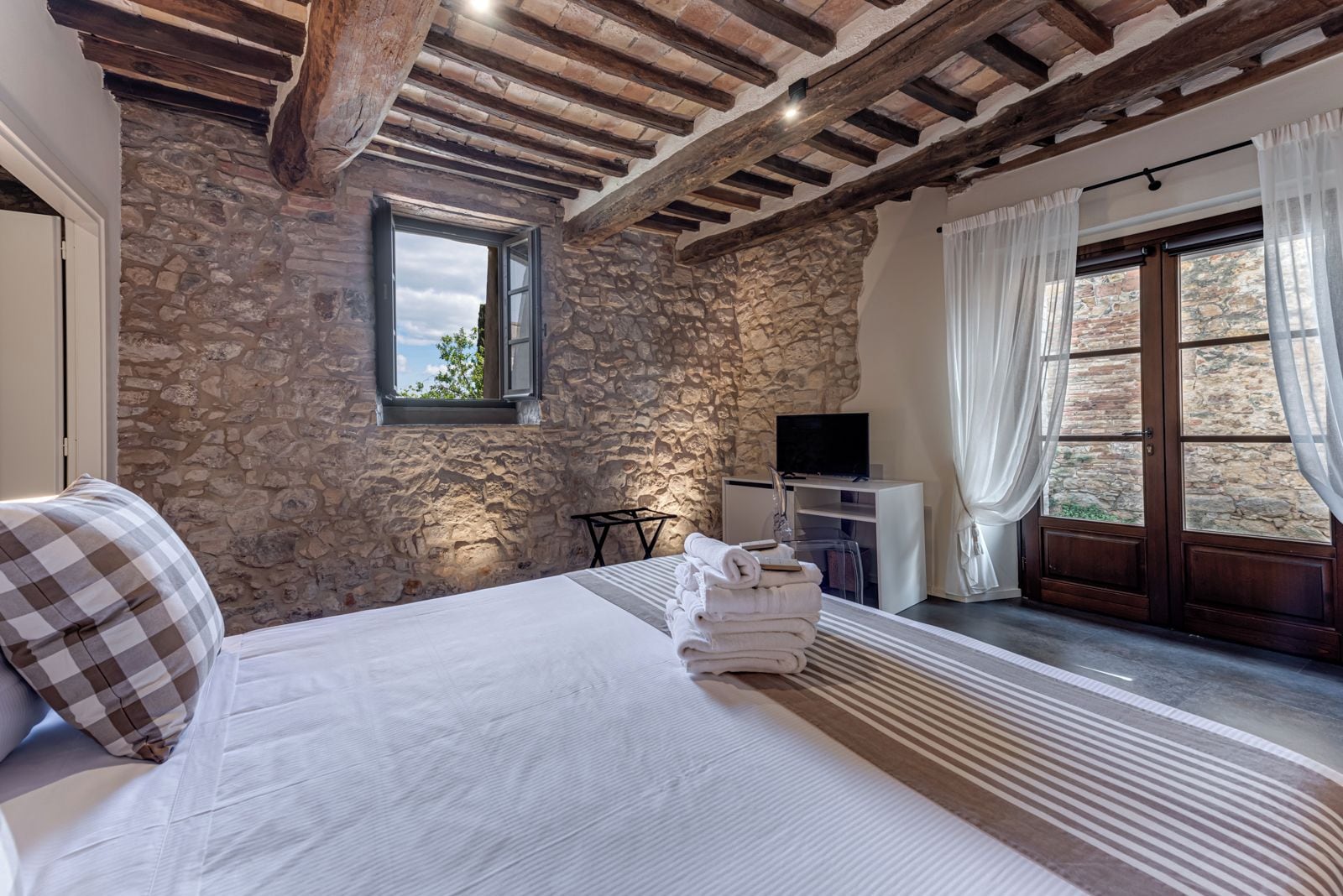 A bedroom with stone walls, a bed with white linens, a television, and a window with curtains.