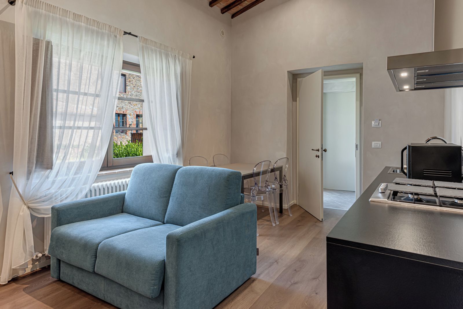 Un salon moderne comprenant un canapé, une table à manger avec des chaises, un coin cuisine avec une cuisinière, et une vue sur un immeuble à travers des rideaux transparents.