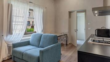 Un salon moderne comprenant un canapé, une table à manger avec des chaises, un coin cuisine avec une cuisinière, et une vue sur un immeuble à travers des rideaux transparents.