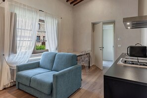 Una sala moderna con un sofá, una mesa de comedor con sillas, una zona de cocina con estufa y vistas a un edificio a través de cortinas translúcidas.