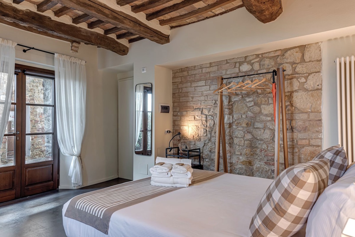 Quarto com parede de pedra, vigas de madeira, cama com roupa de cama branca, cabideiro de madeira e janela com cortinas.