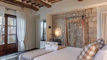 A bedroom with a stone wall, wooden beams, a bed with white linens, a wooden hanger rack, and a window with curtains.