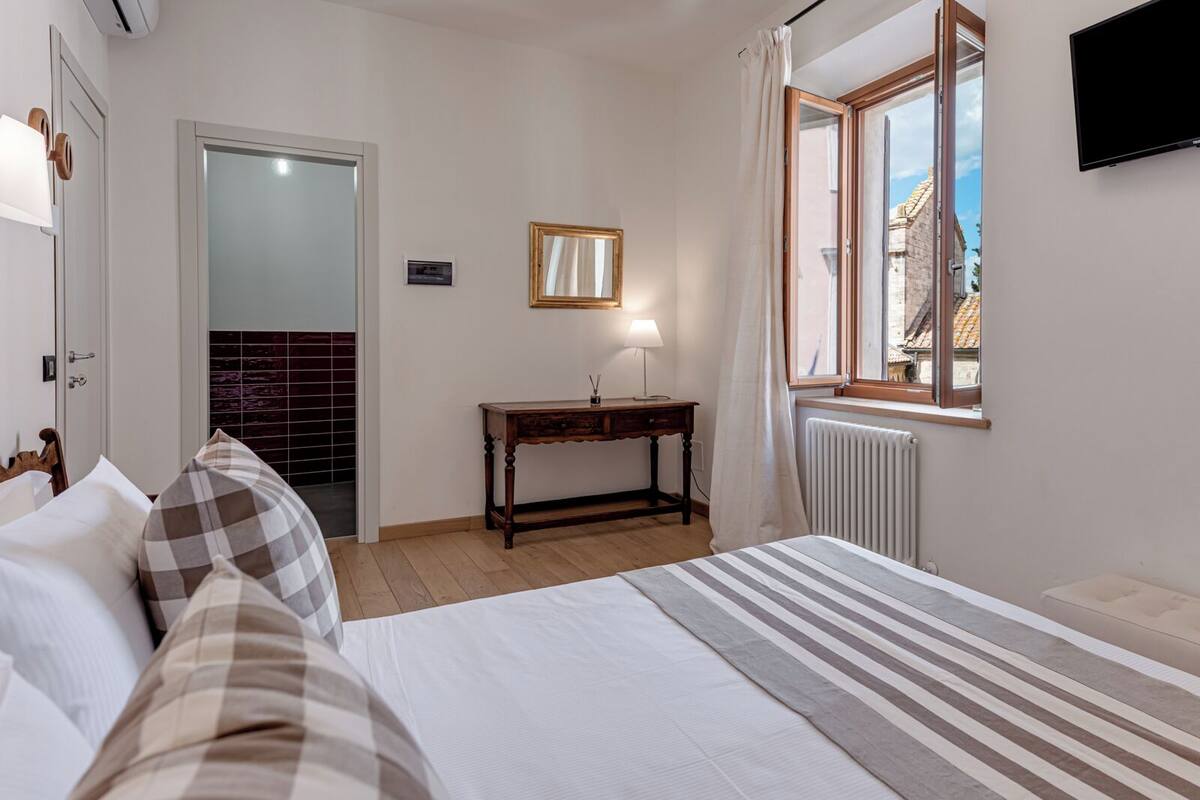 Una camera da letto con un letto, un comodino, uno specchio, una finestra con tende e vista sugli edifici esterni.