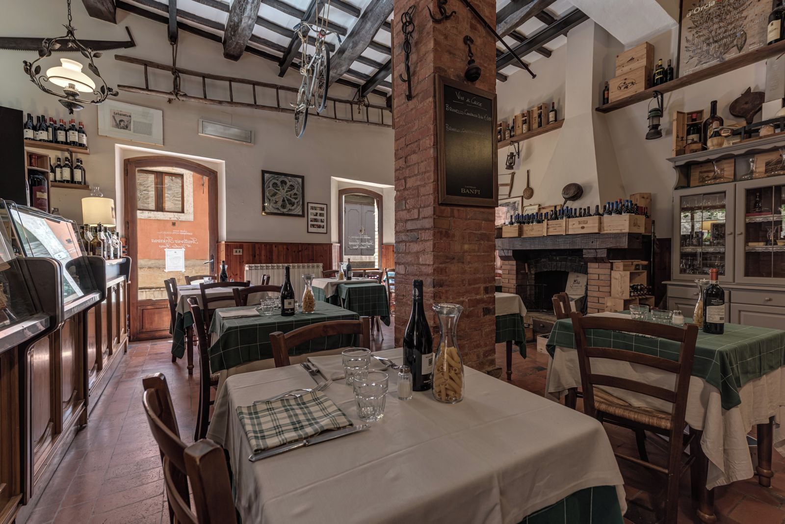 Un restaurant aux murs de briques, aux poutres apparentes et où l’on peut admirer une variété de bouteilles de vin exposées sur des étagères. Les tables sont dressées avec des verres à vin et des assiettes.