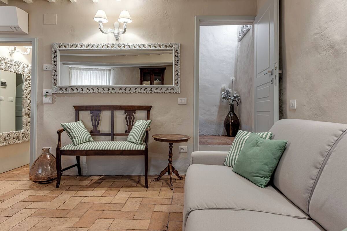Un soggiorno con un divano, una panca di legno con cuscini a righe, un tavolino rotondo e un grande specchio con cornice decorata.