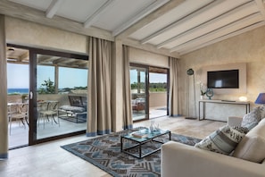 A spacious living area with a large sliding glass door leading to an outdoor patio, a comfortable sofa, a coffee table, and a flat-screen TV mounted on the wall. - Baglioni Resort Sardinia (San Teodoro)