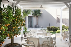 Un espace extérieur aménagé pour les repas, avec des tables et des chaises blanches, un arbre en pot orné d’oranges, et un escalier en arrière-plan.