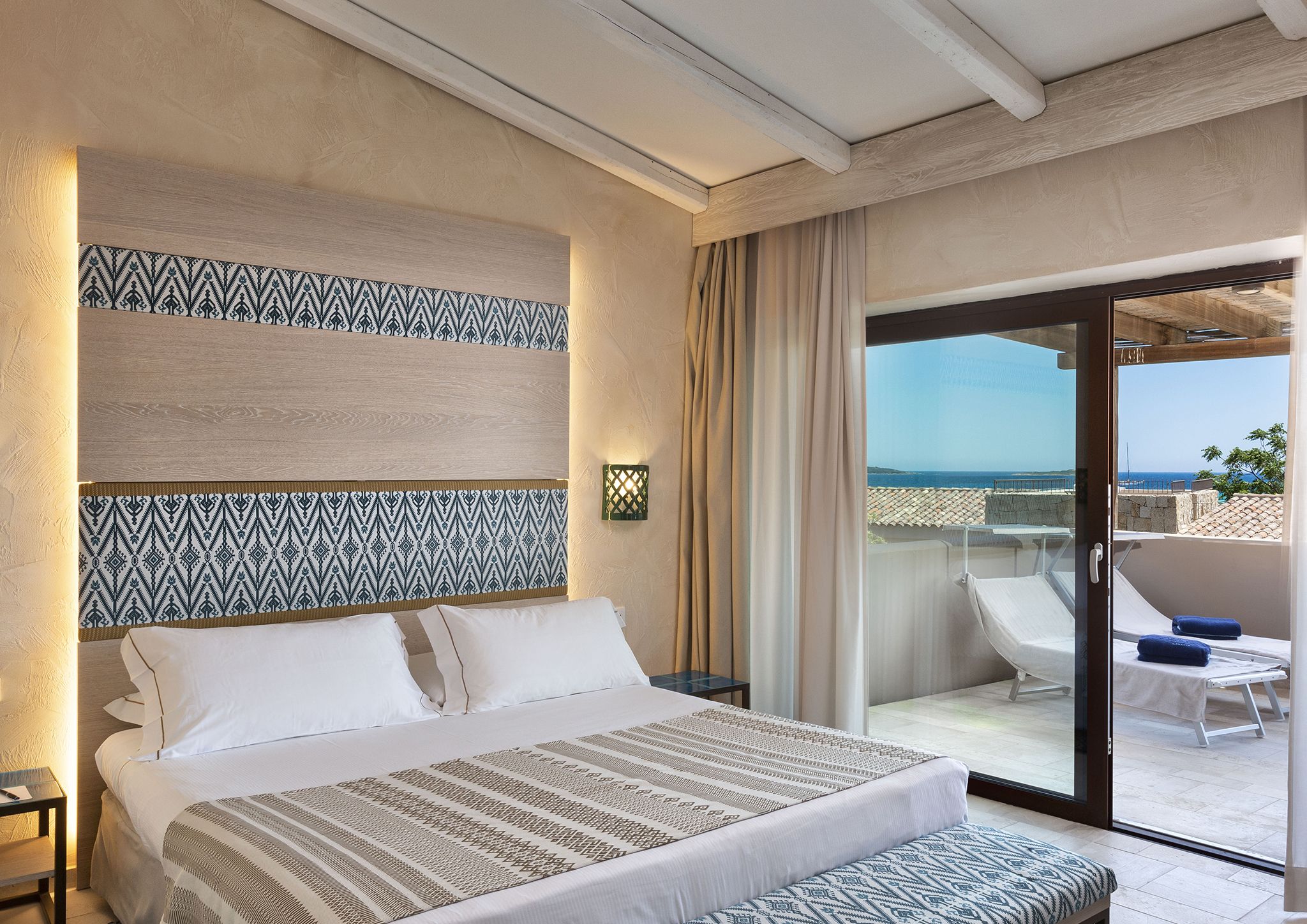 A hotel room with a bed, a nightstand, a patterned headboard, a large window with a view of the sea, and a balcony with a chair and table.