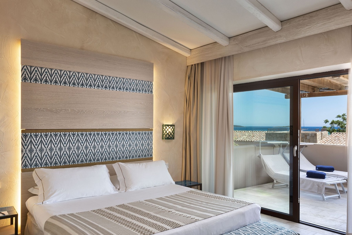 A hotel room with a bed, a nightstand, a patterned headboard, a large window with a view of the sea, and a balcony with a chair and table.