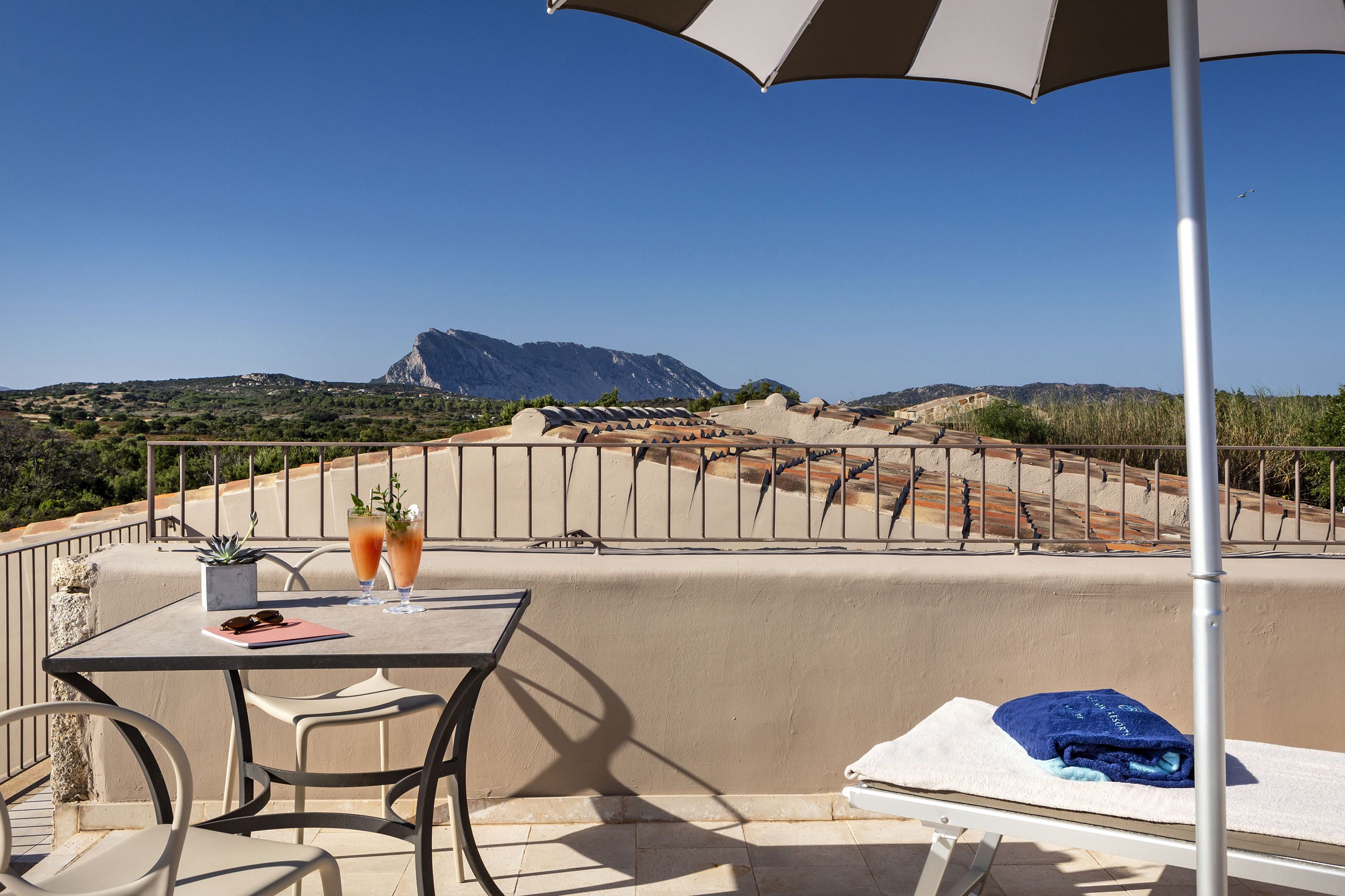 A terrace with a table, chairs, a sun umbrella, a towel, and a view of mountains.