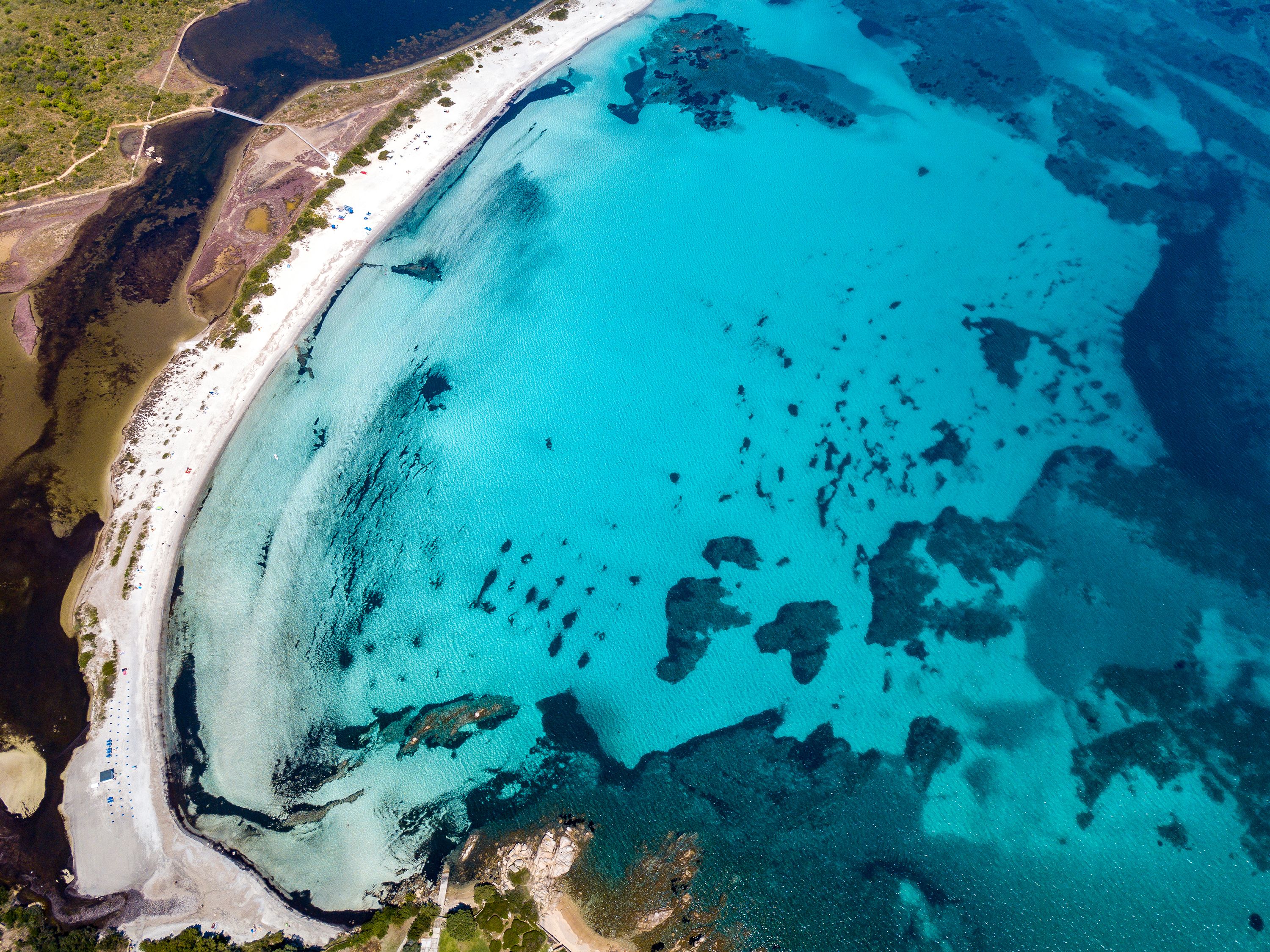 Ett flygbildsbild av en strand med kristallklart turkosfärgat vatten, en sandstrand och här och var lite växtlighet.