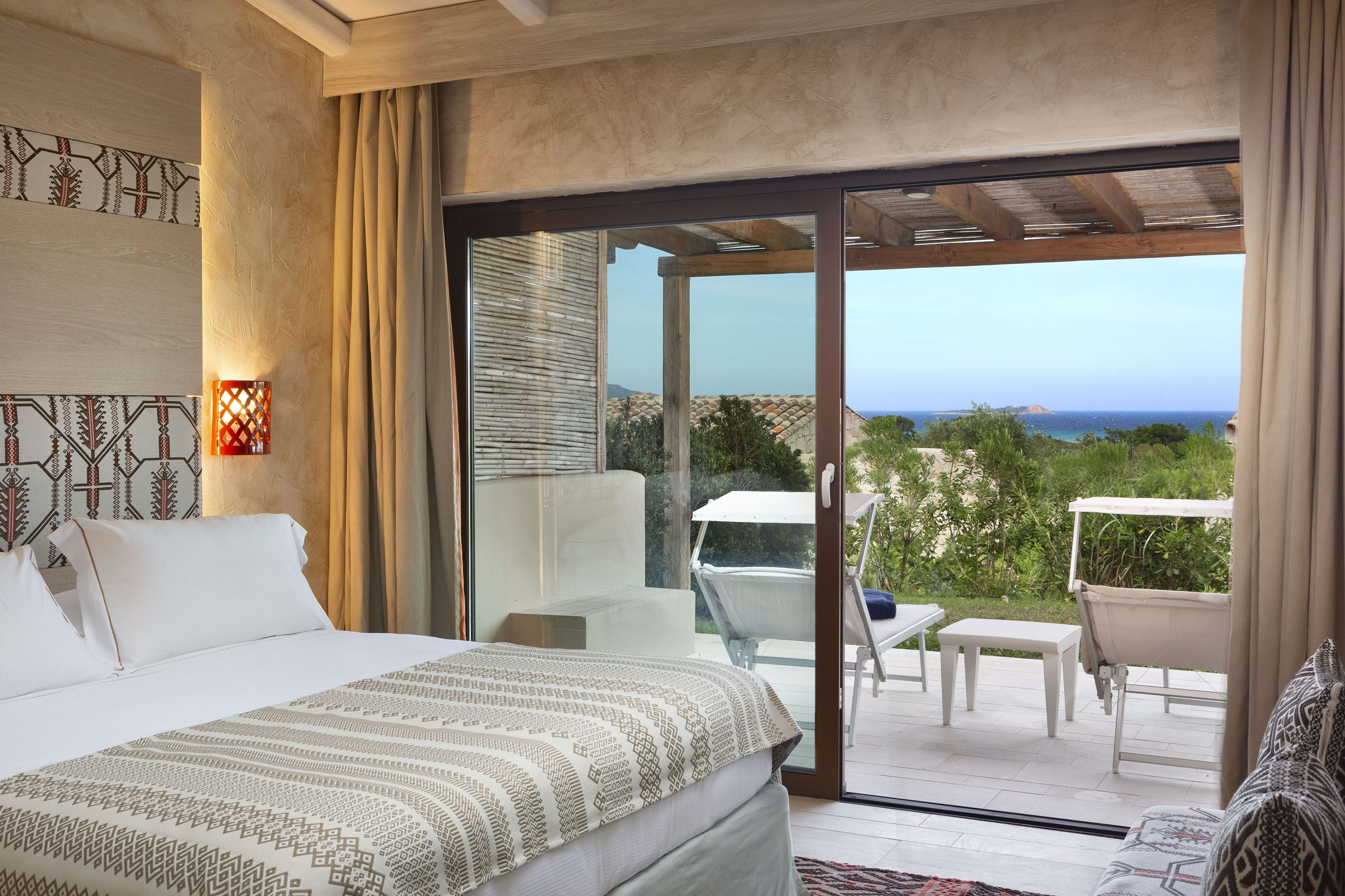 Une chambre d’hôtel avec un lit, une petite table et une chaise. Une porte-fenêtre coulissante donne accès à un espace extérieur d’où l’on peut admirer l’océan.