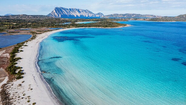 Un paisaje costero con una playa, aguas azules cristalinas y una montaña al fondo.