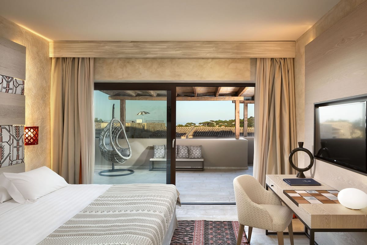 A modern hotel room with a bed, desk, chair, TV, and a view of the cityscape.